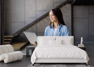 Attractive smiling young brunette woman using laptop computer while standing in a stylish living room, Wall mural